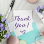 A beautifully arranged thank you card with lavender flowers, macarons, and gift package on a marble desk.