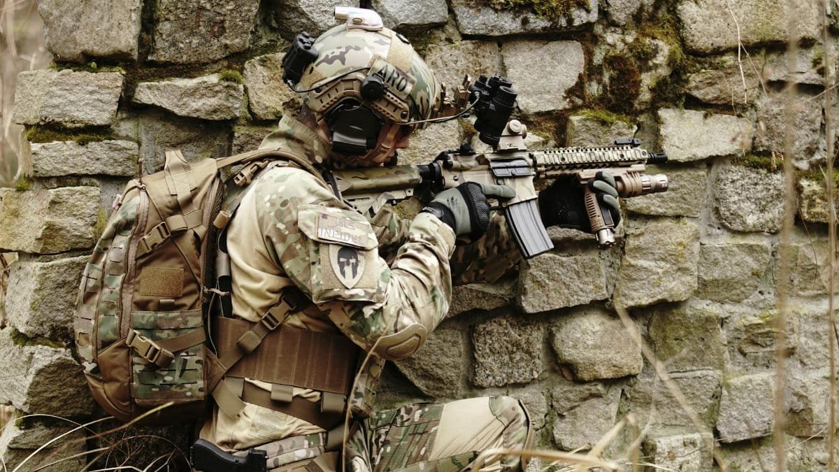Soldier in camouflage gear aims a rifle while hiding along a stone wall outdoors.