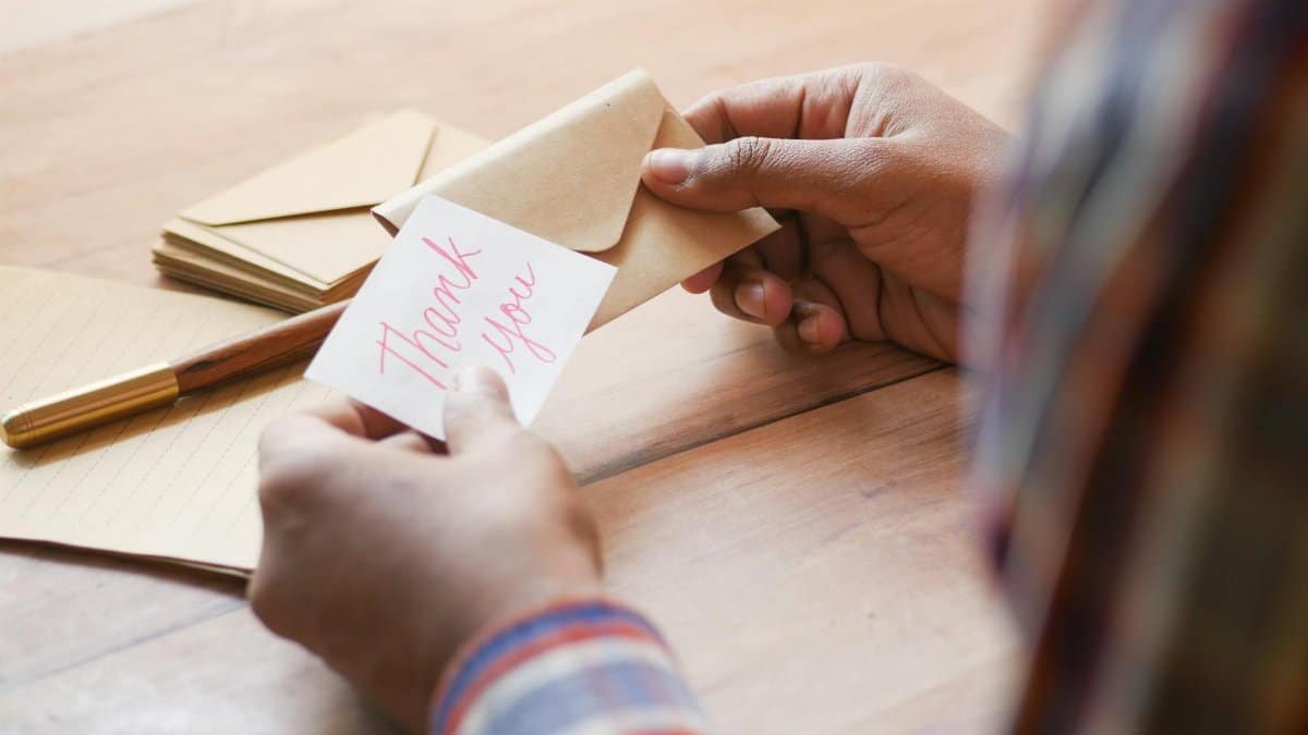Close-up of hands holding a thank you note in an envelope on a table.