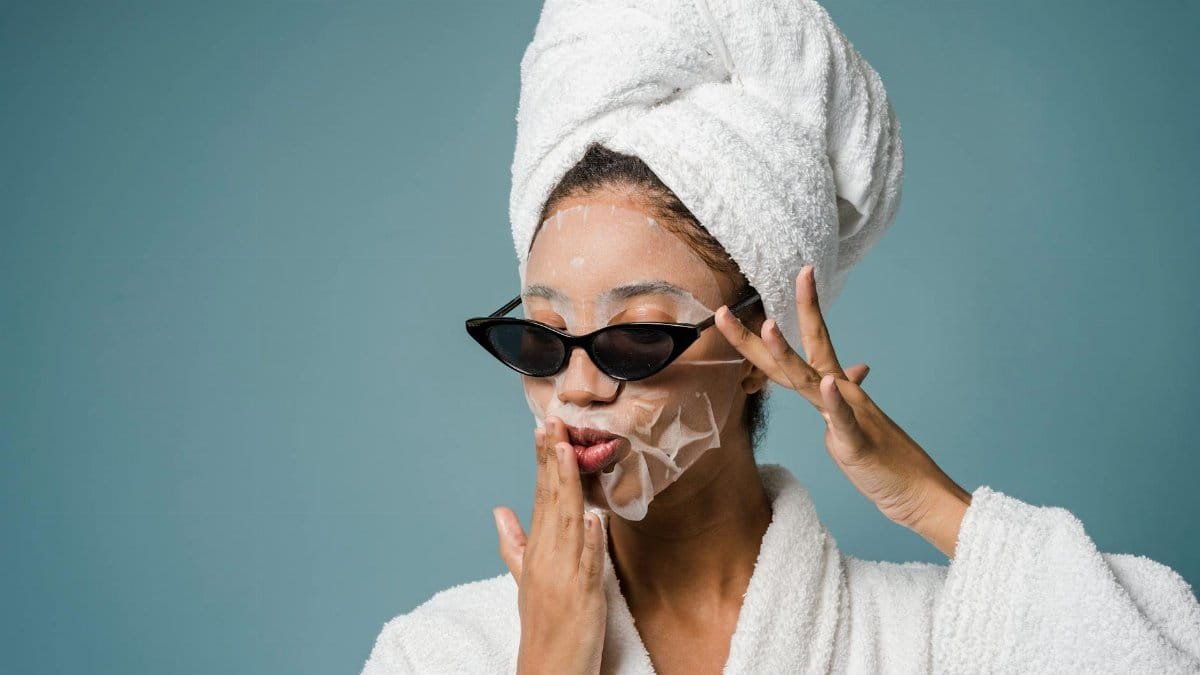Calm African American woman with moisturizing sheet mask in sunglasses and white towel turban on head touching dace against blue background in studio
