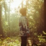 A person stands thoughtfully in a sunlit forest surrounded by tall trees and glowing foliage.