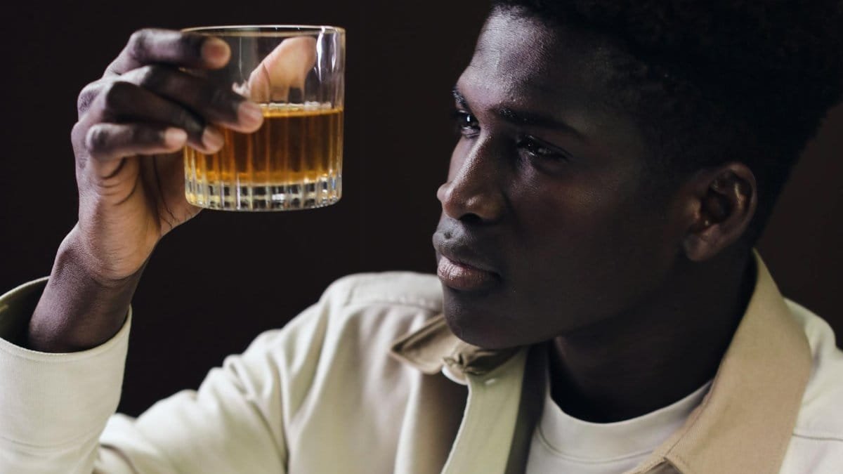 An adult man with curly hair carefully examines a glass of whiskey, captured in a thoughtful close-up portrait indoors.