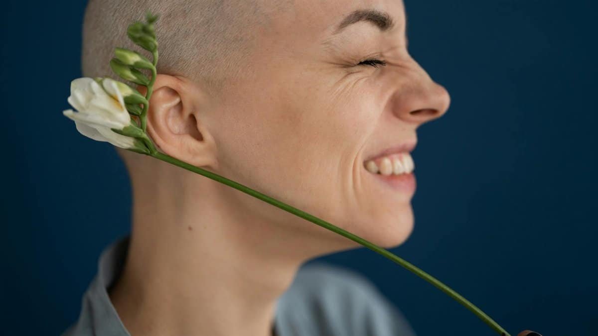 Side view of crop cheerful female patient with blossoming flower representing scar on head concept after brain tumor surgery on blue background