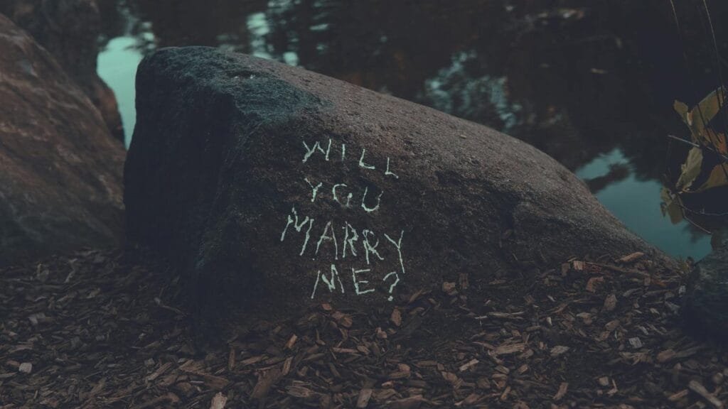 Chalk-written marriage proposal on a rock by a lake in a serene forest setting.