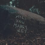 Chalk-written marriage proposal on a rock by a lake in a serene forest setting.