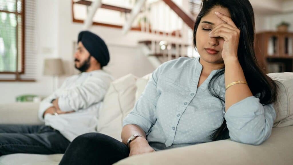 A young couple sitting on a couch indoors, showing signs of stress and frustration.