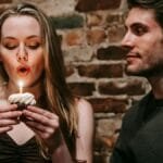 Young couple in elegant outfits in restaurant while blowing in candle on small cupcake and celebrating birthday near brick wall