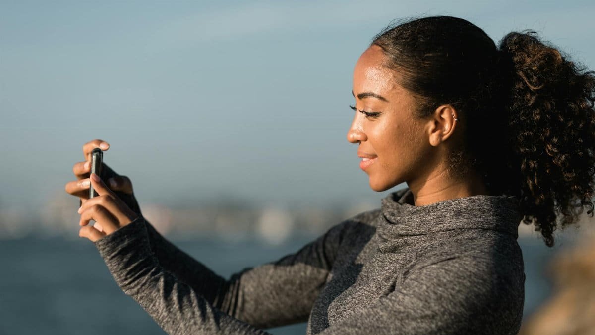 A young African American woman taking a selfie outdoors by the water on a sunny day.