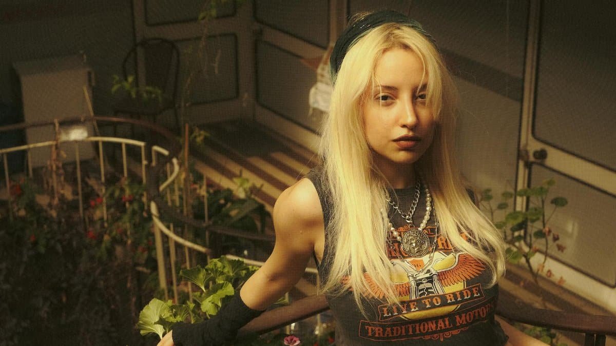 Blonde woman posing in an indoor garden wearing a biker-themed top, exuding a modern and edgy vibe.