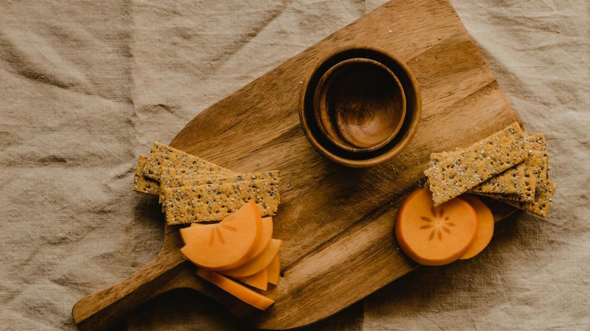 Flat lay of wood board with sliced persimmon, crackers, and bowls for a rustic vibe.