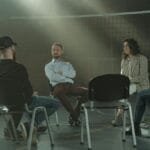 A group therapy session with six adults seated in a circle, discussing support and mental health.