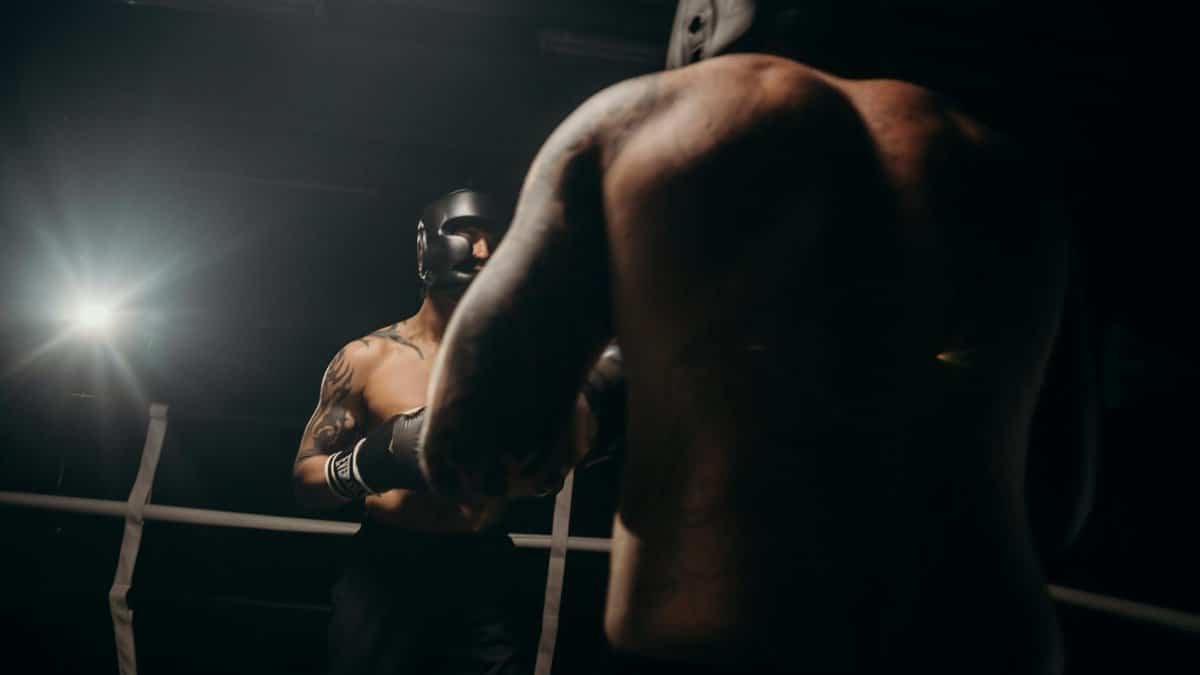 Two boxers sparring in a dimly lit gym, wearing head guards for protection.