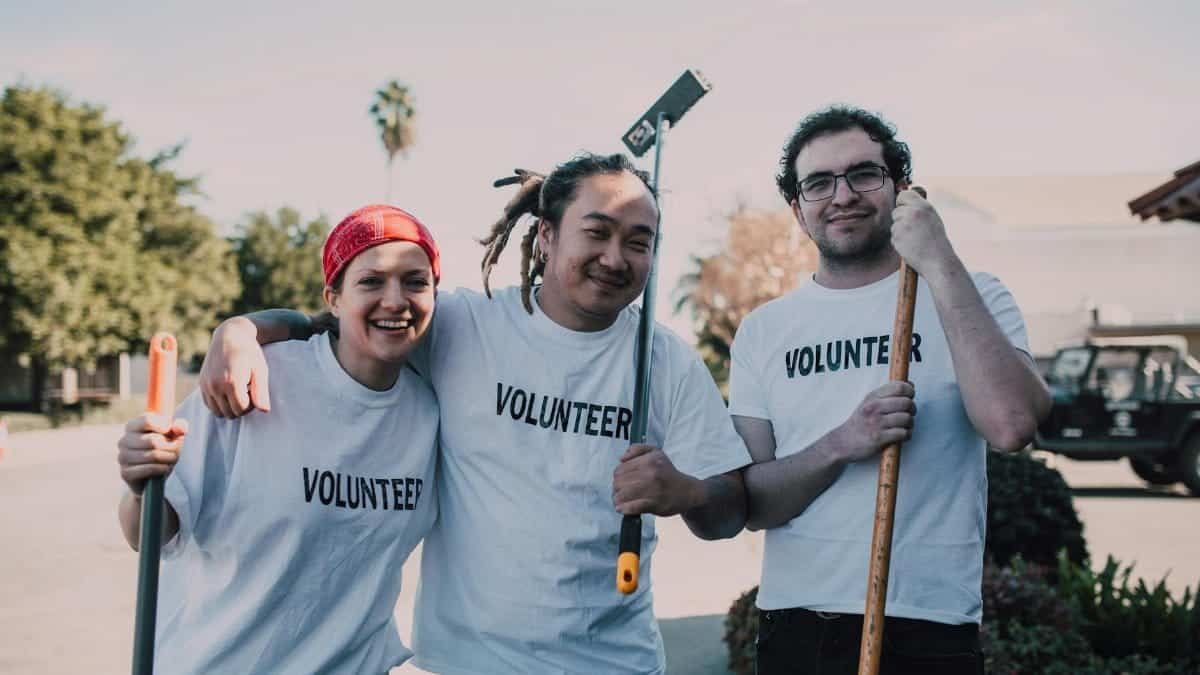 Three diverse volunteers smiling and holding cleaning tools during a community service event.