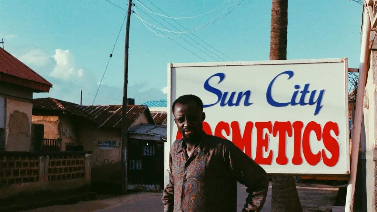 Man with patterned shirt standing by Sun City Cosmetics sign in urban setting.