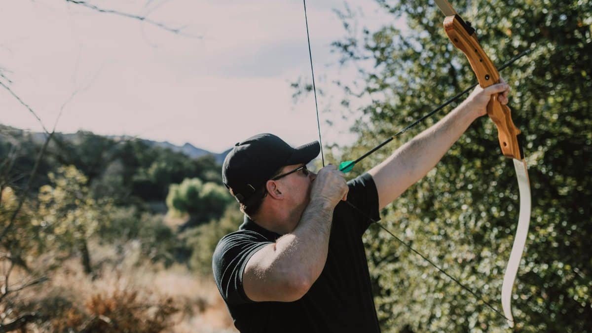 An adult man draws his recurve bow aiming for a target during an outdoor archery practice session.
