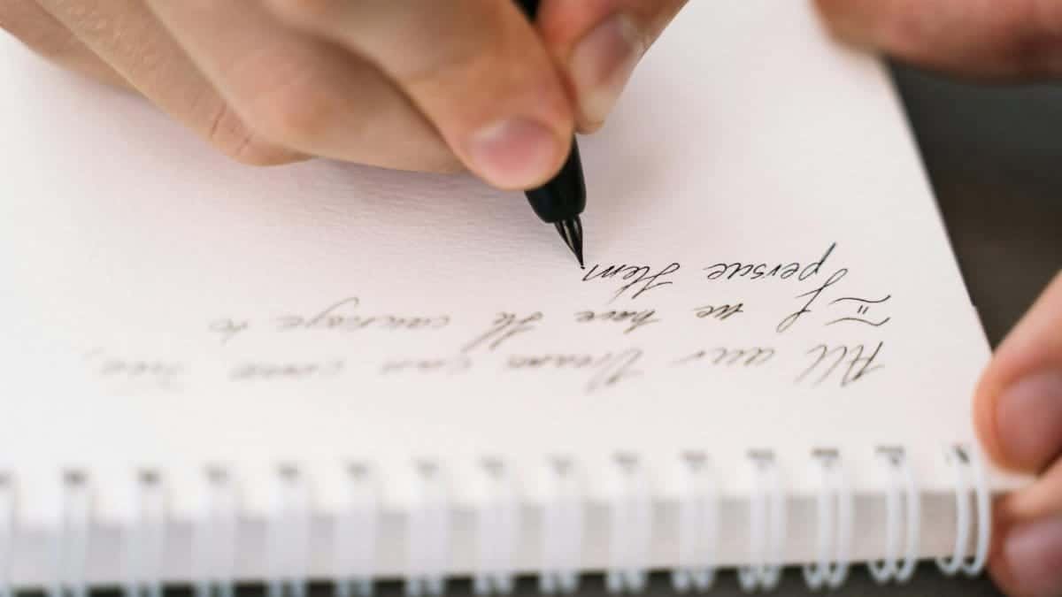 A person writes in a spiral notebook with elegant handwriting, using a pen.