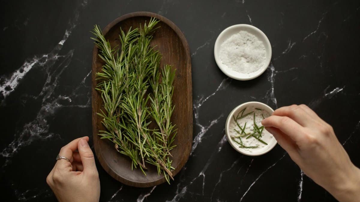 Top view of unrecognizable woman adding fresh twigs of rosemary in bowl with salt for organic scrub placed on black marble table