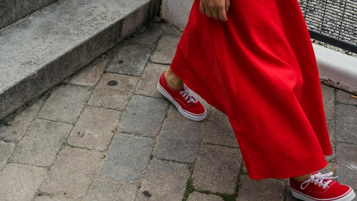 Woman wearing a red dress and sneakers walking on a cobblestone sidewalk.