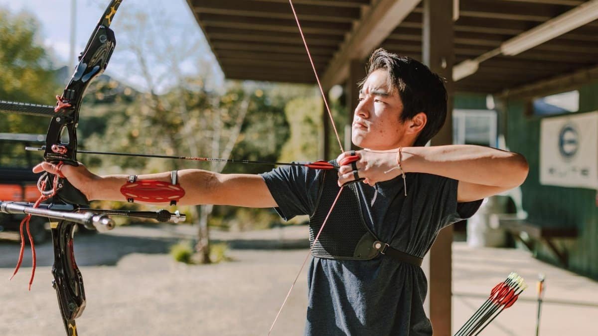 Asian man practicing archery with a recurve bow outdoors on a sunny day, focused and aiming.