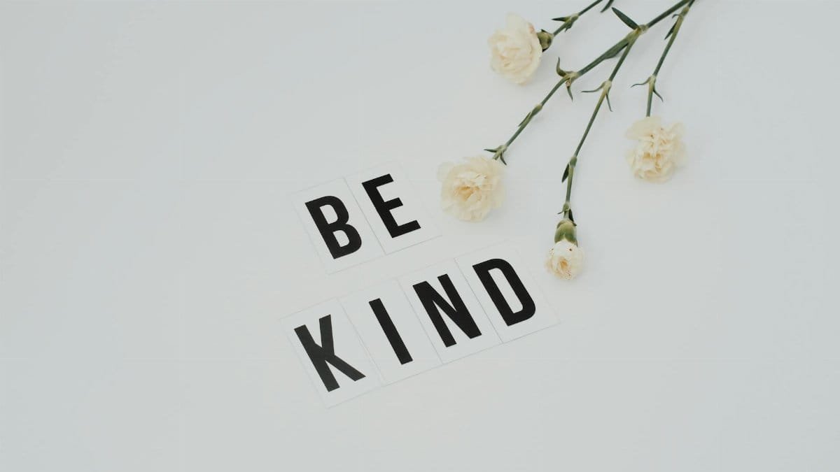 Minimalist image of 'Be Kind' text with white flowers on a light background, promoting kindness.