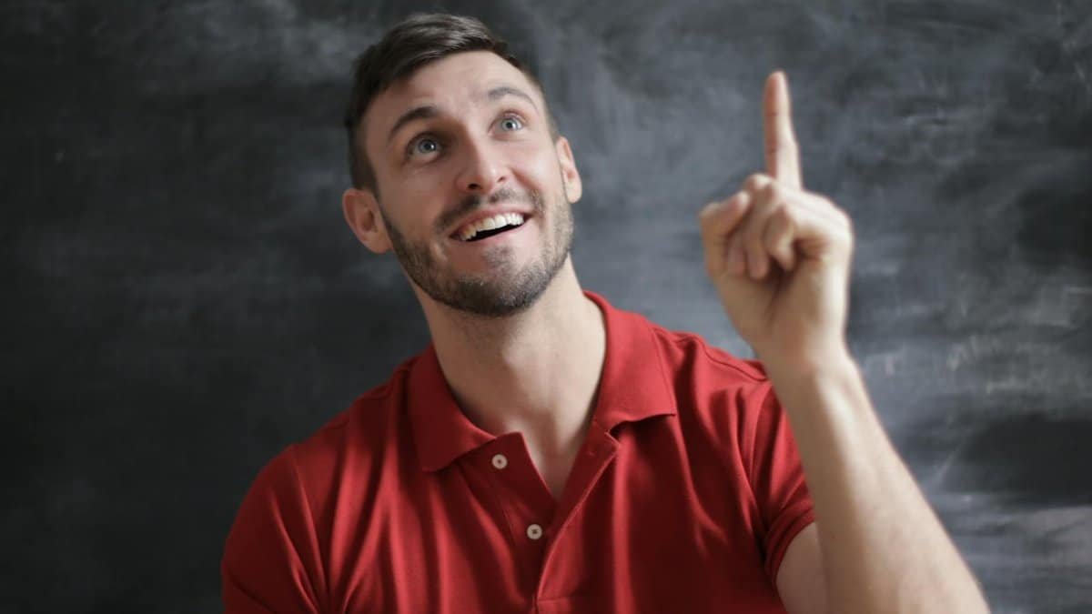 Happy young man in a red polo shirt smiling and pointing upwards in front of a blackboard.