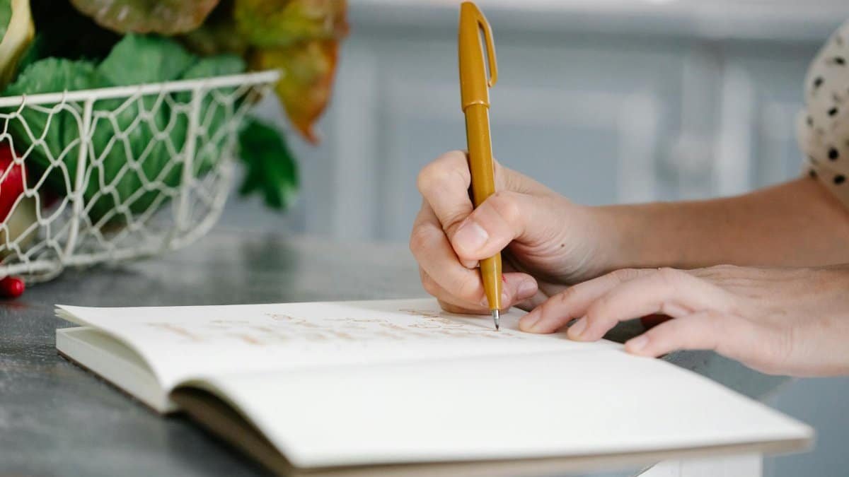 A woman writes in a notebook on a countertop near a basket of fresh produce.