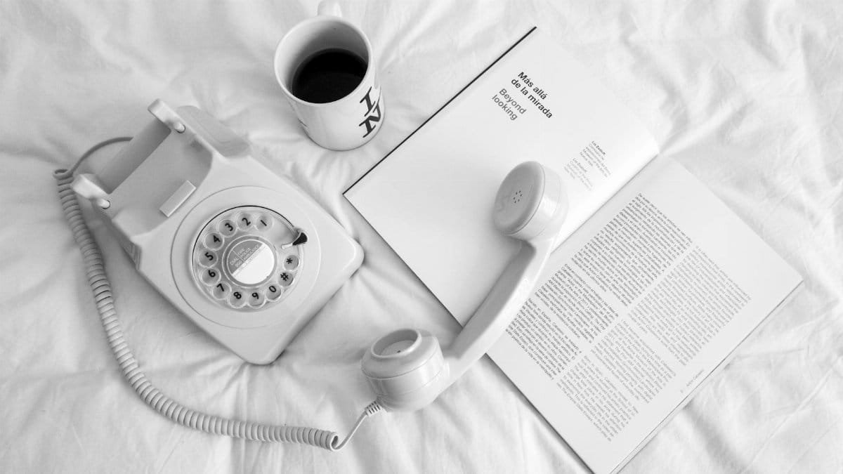 A cozy scene with a vintage rotary phone, coffee, and book on a white bed.