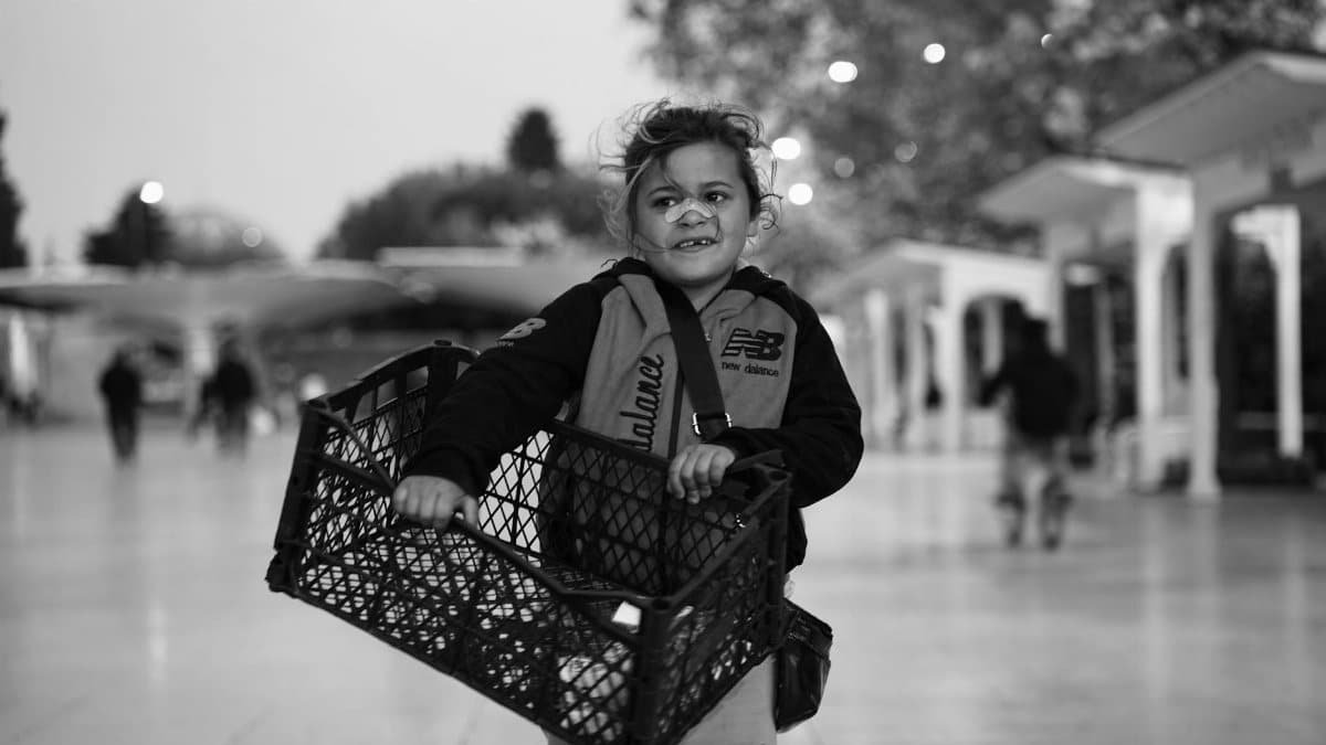 Black and white photo of a child carrying a crate, capturing a candid street moment outdoors.