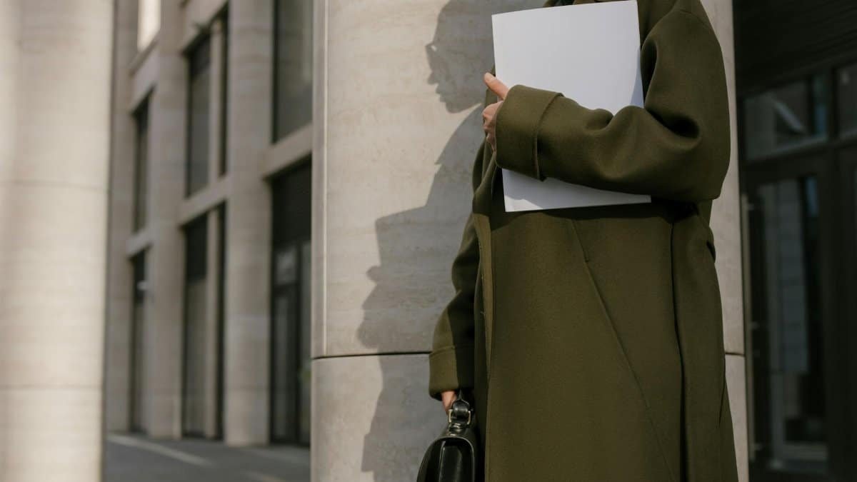 Woman in a green coat holding documents outside a modern office building.