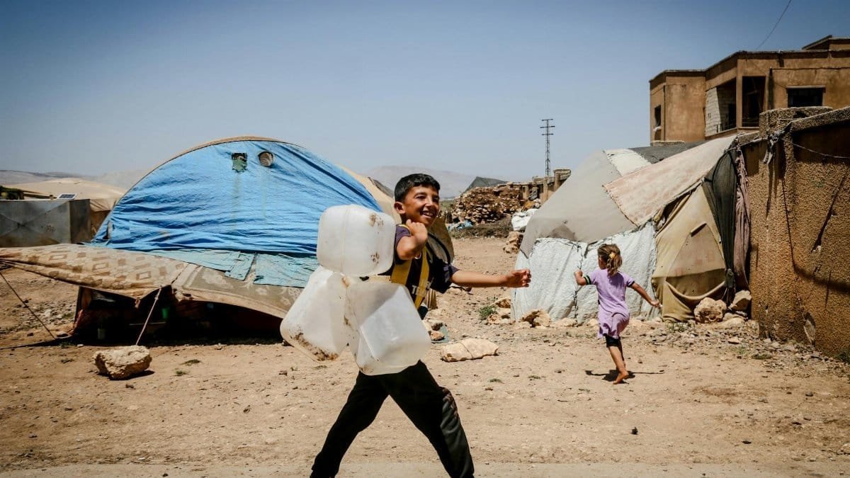 Children in Idlib refugee camp carrying water jugs, smiling and walking.