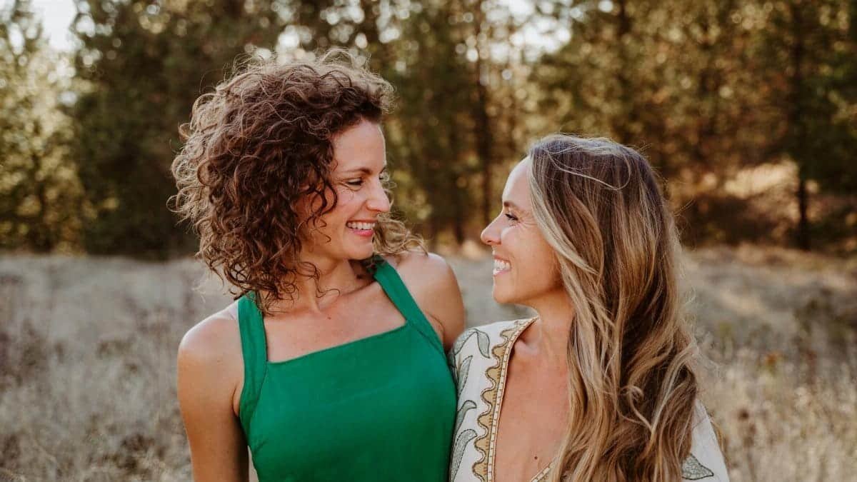 Two women smiling and embracing outdoors in a sunlit meadow, expressing joy and connection.