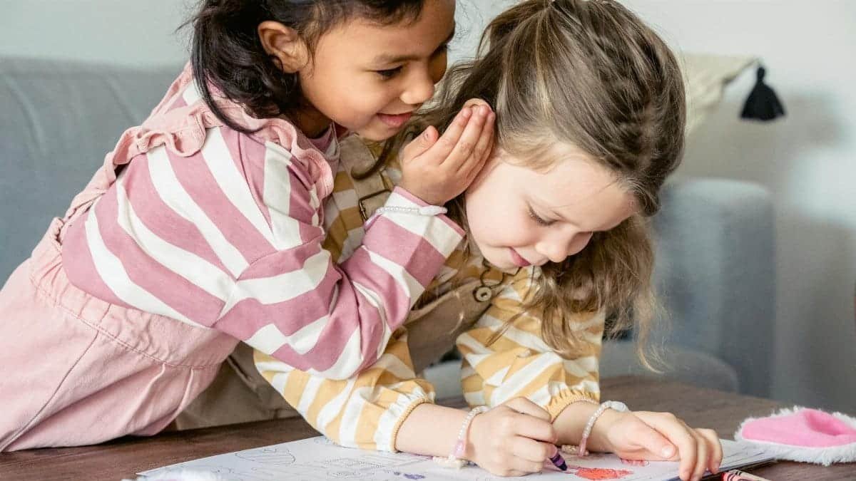 Two young girls whisper secrets while coloring together at a table.