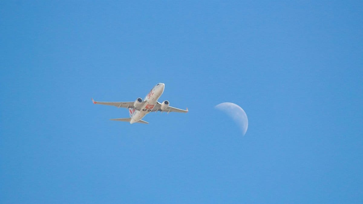 Airplane flying through a clear blue sky with the moon visible.