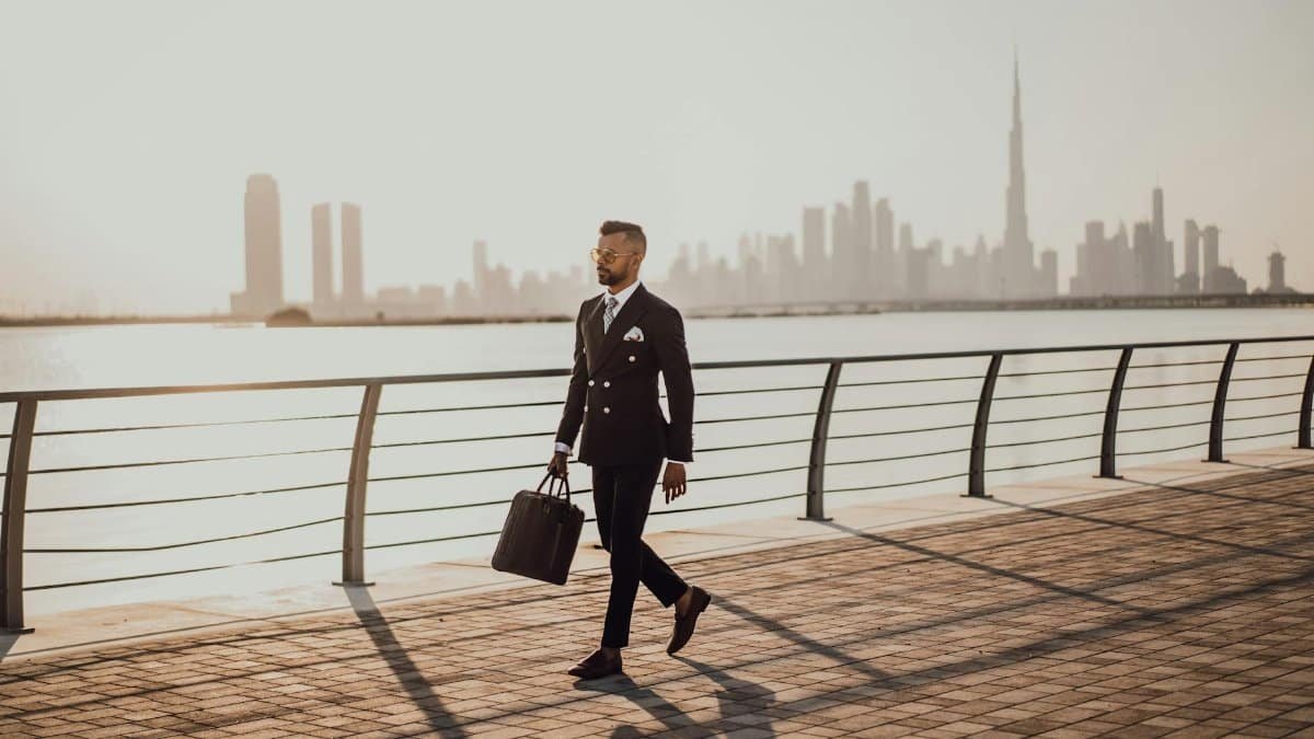 Professional man in formal suit walking by Dubai waterfront with city skyline and Burj Khalifa in view.