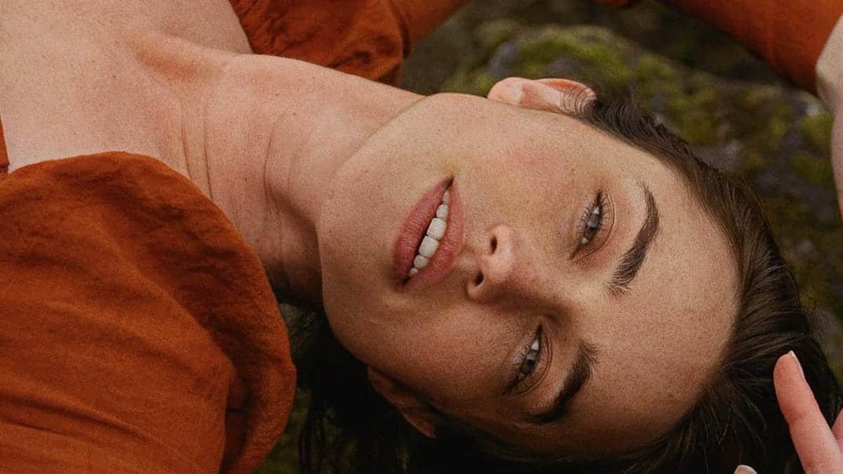 A serene portrait of a young woman in an orange blouse lying outdoors on moss-covered ground.