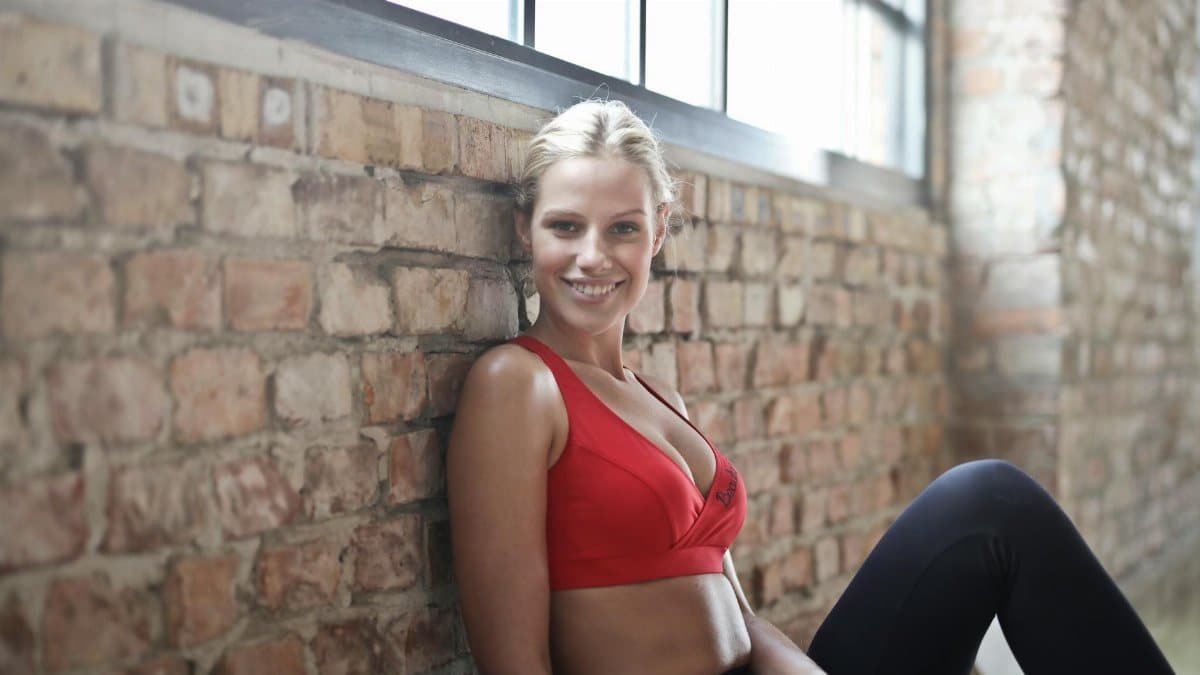 A fit woman in a red sports bra smiling while sitting against a brick wall after a workout.