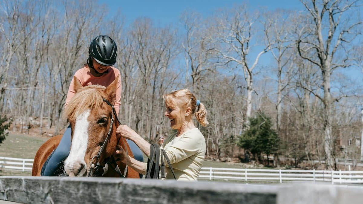A woman giving a horse riding lesson to a child in a sunny outdoor arena, emphasizing safety and fun.