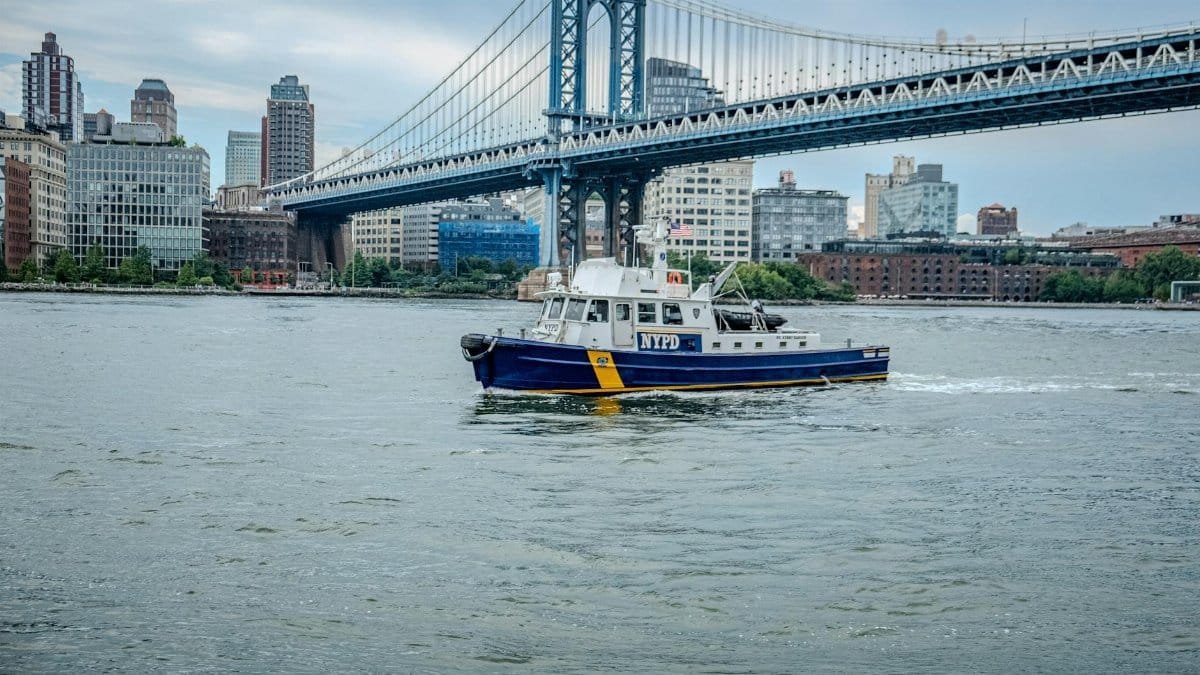 NYPD boat cruising near the iconic Manhattan Bridge with New York City skyline in the background.