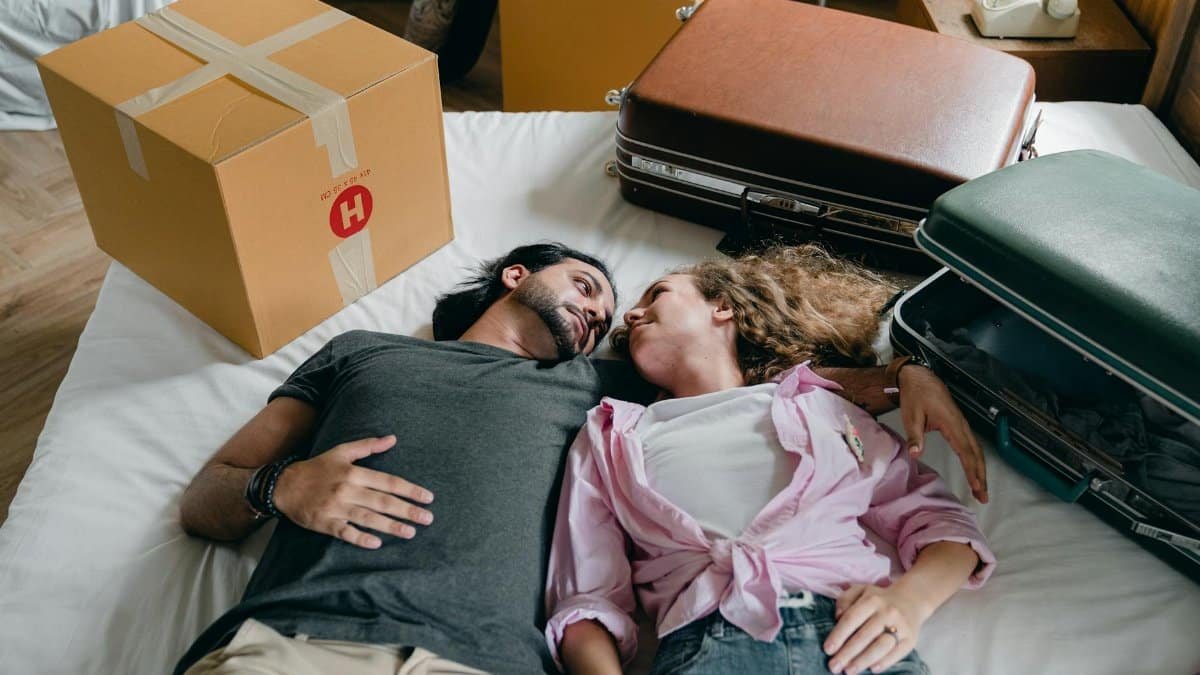 From above of young ethnic bearded man and woman with curly hair looking at each other and lying on bed among suitcases and cardboard boxes with stuff while moving in new apartment together