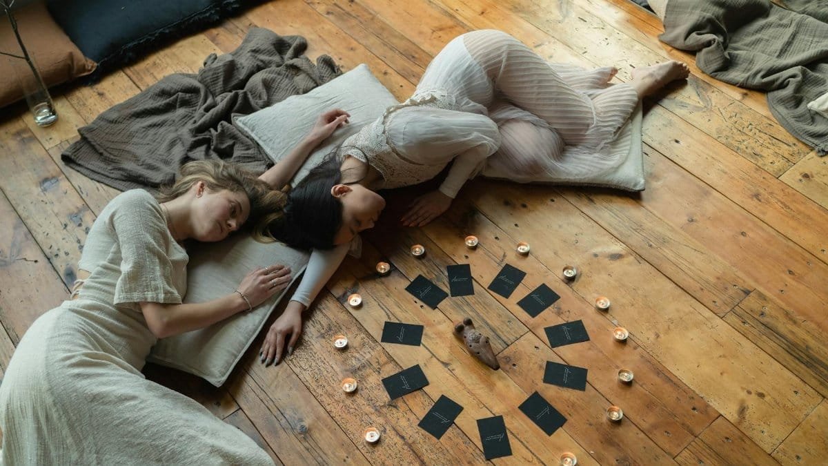 Two women lying on wooden floor, surrounded by candles and tarot cards, in a spiritual setting.