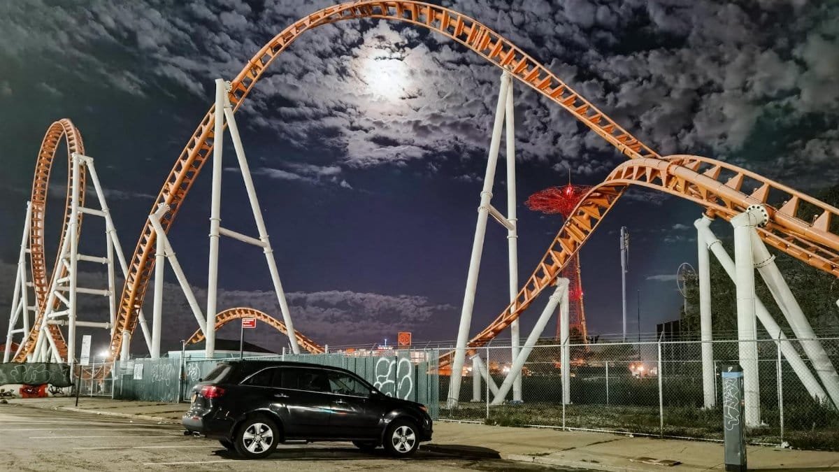 A dramatic nighttime shot of a rollercoaster in New York City with a car parked nearby under a bright moon.