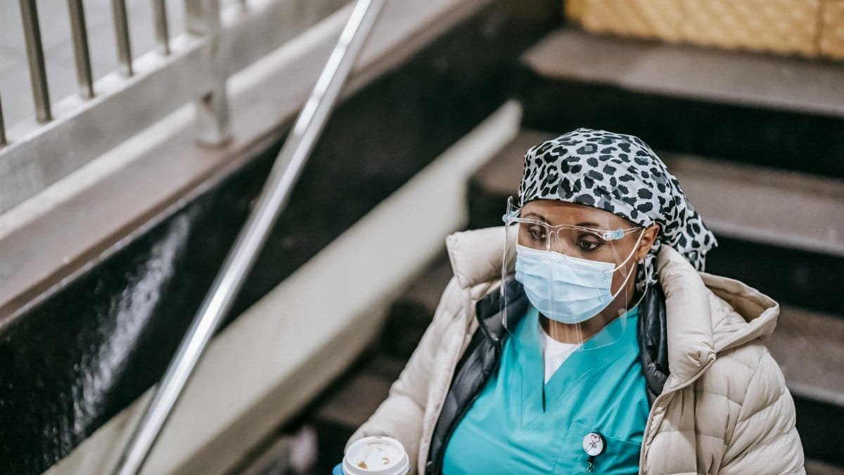 A healthcare worker wearing a mask and gloves descends stairs with a coffee cup.