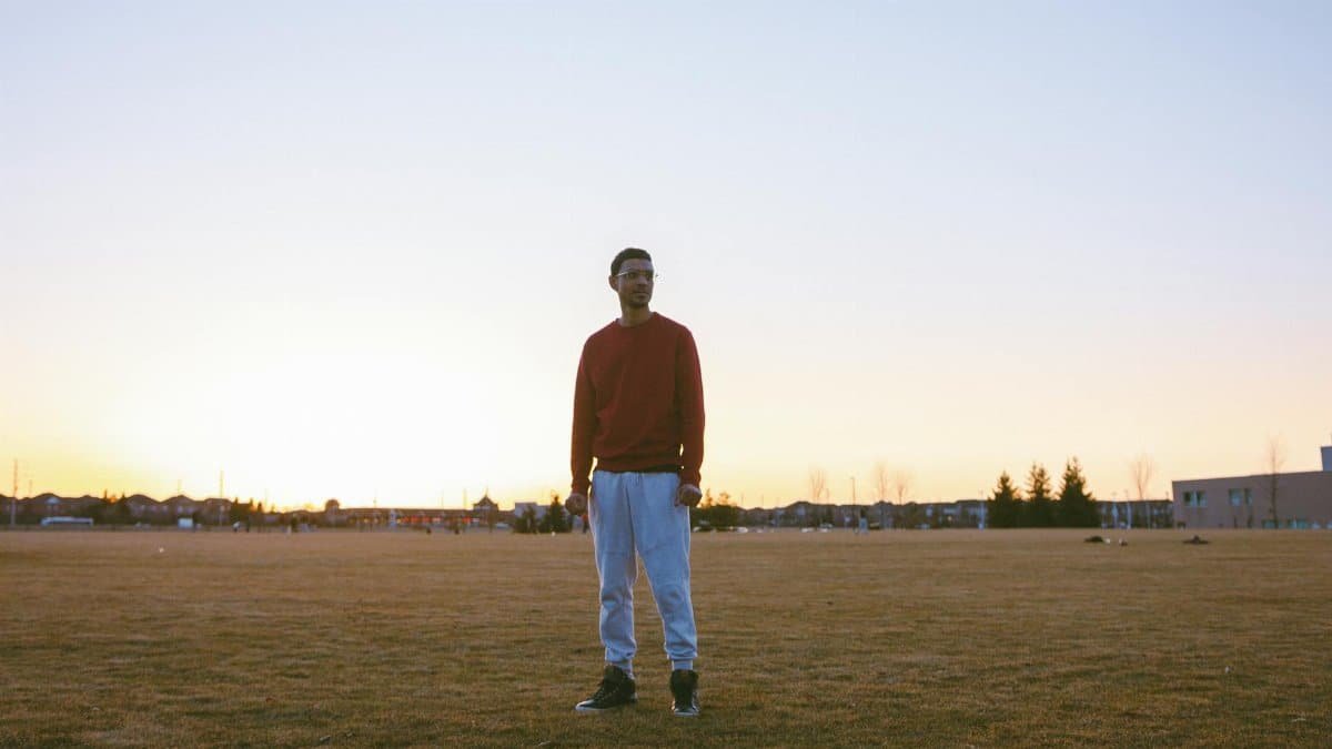 A solitary man stands in an open field during sunrise, wearing a red sweater.