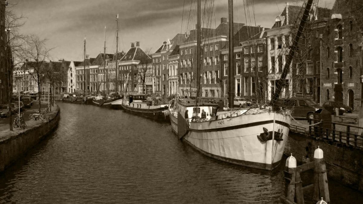 Sepia-toned image of a canal with historical sailboats and classic architecture.