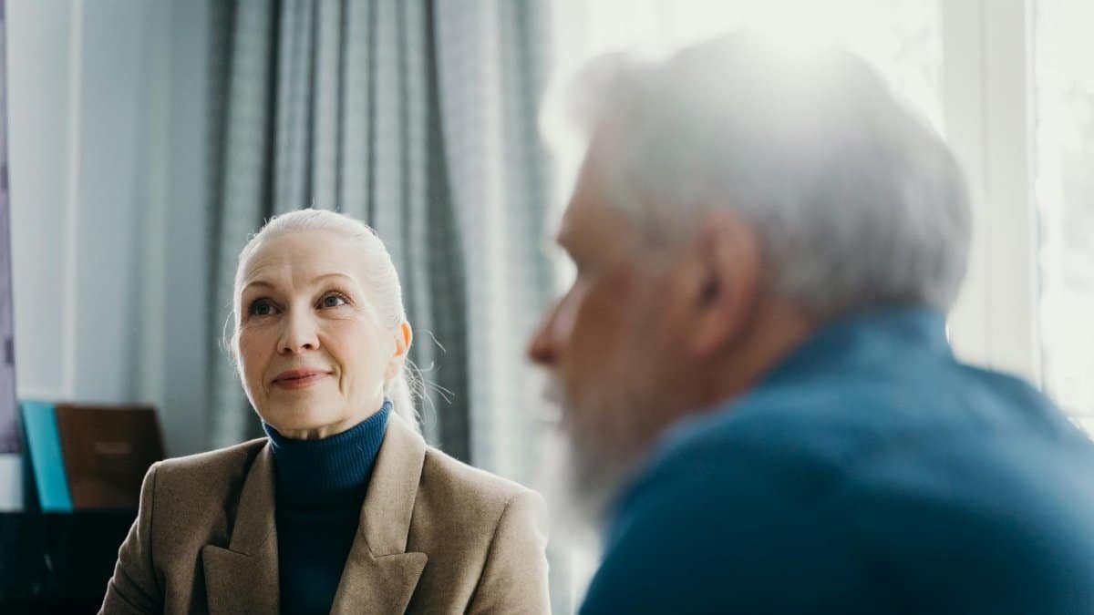 Elderly man and woman engaged in a warm conversation, sunlight streaming through the window.
