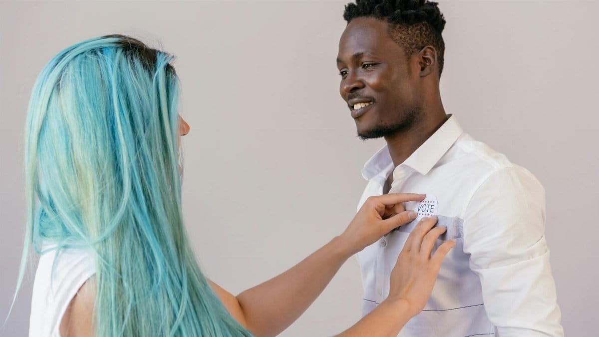 A woman pins a voting badge on a smiling man's shirt, promoting civic engagement.