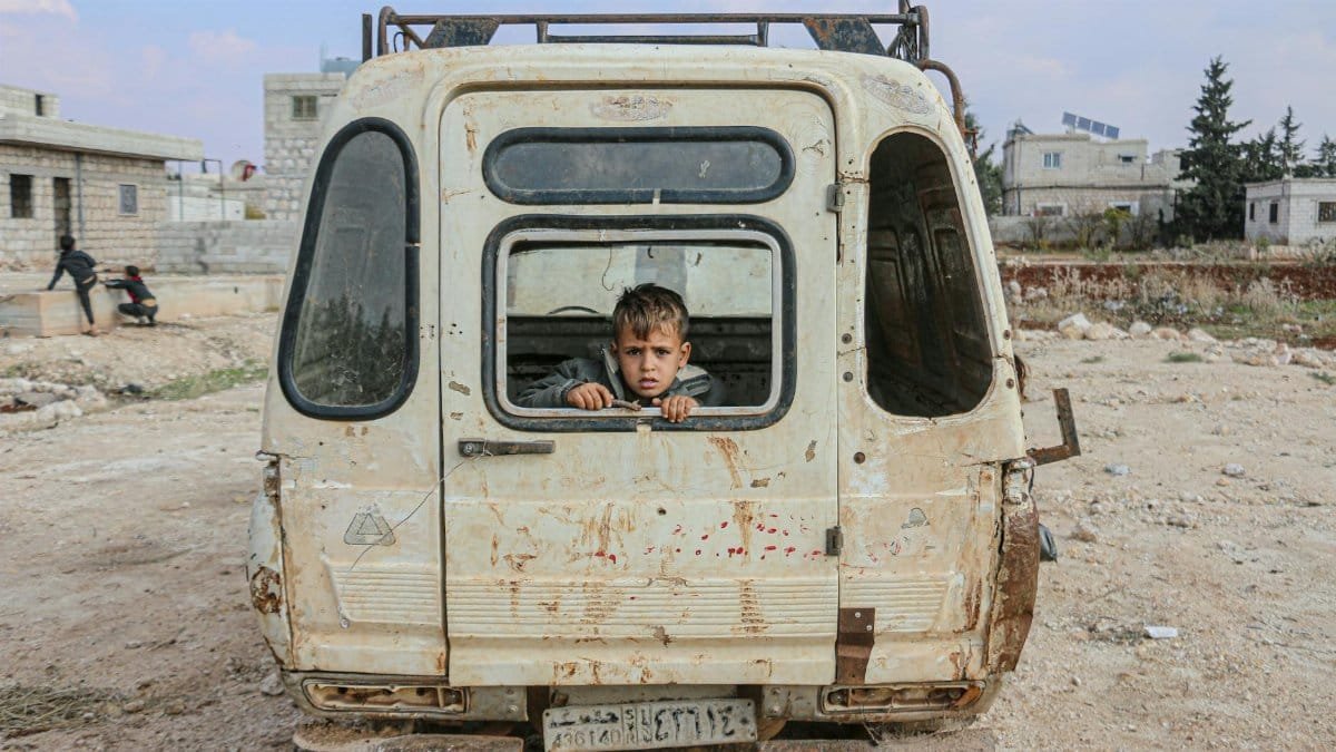 A young boy peers from an abandoned van in the desolate streets of Idlib, Syria, amidst war devastation.