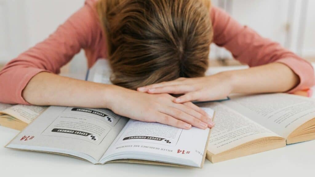 A student overwhelmed by studying falls asleep on open books at a desk, representing academic fatigue.