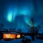 Cabin illuminated under a stunning aurora borealis and starry winter night sky.