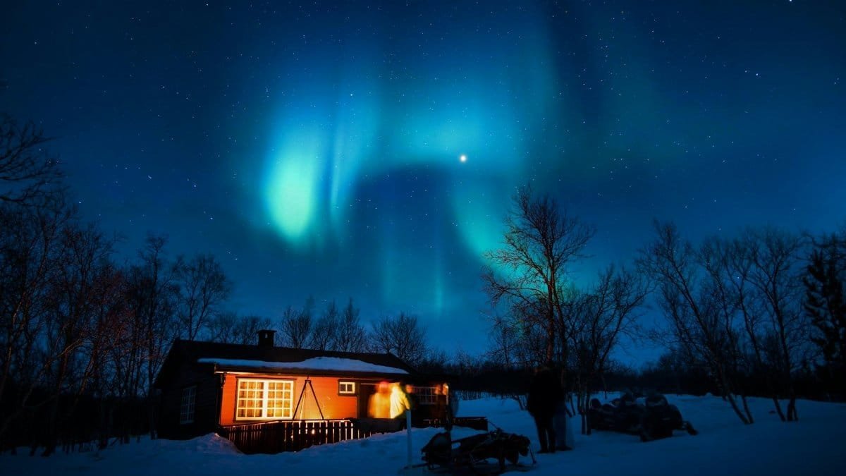 Cabin illuminated under a stunning aurora borealis and starry winter night sky.